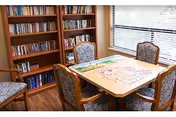 Small community library/activity room with a bookshelf, a table holding a jigsaw puzzle, and four upholstered chairs by a window.