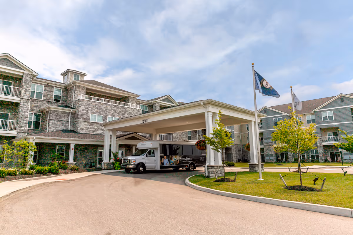 Exterior view of StoryPoint Union senior living facility showing a large stone and siding building with multiple windows and balconies. A covered driveway entrance is visible with a StoryPoint senior living shuttle bus parked underneath. There are two flagpoles with flags and landscaped greenery around the driveway.