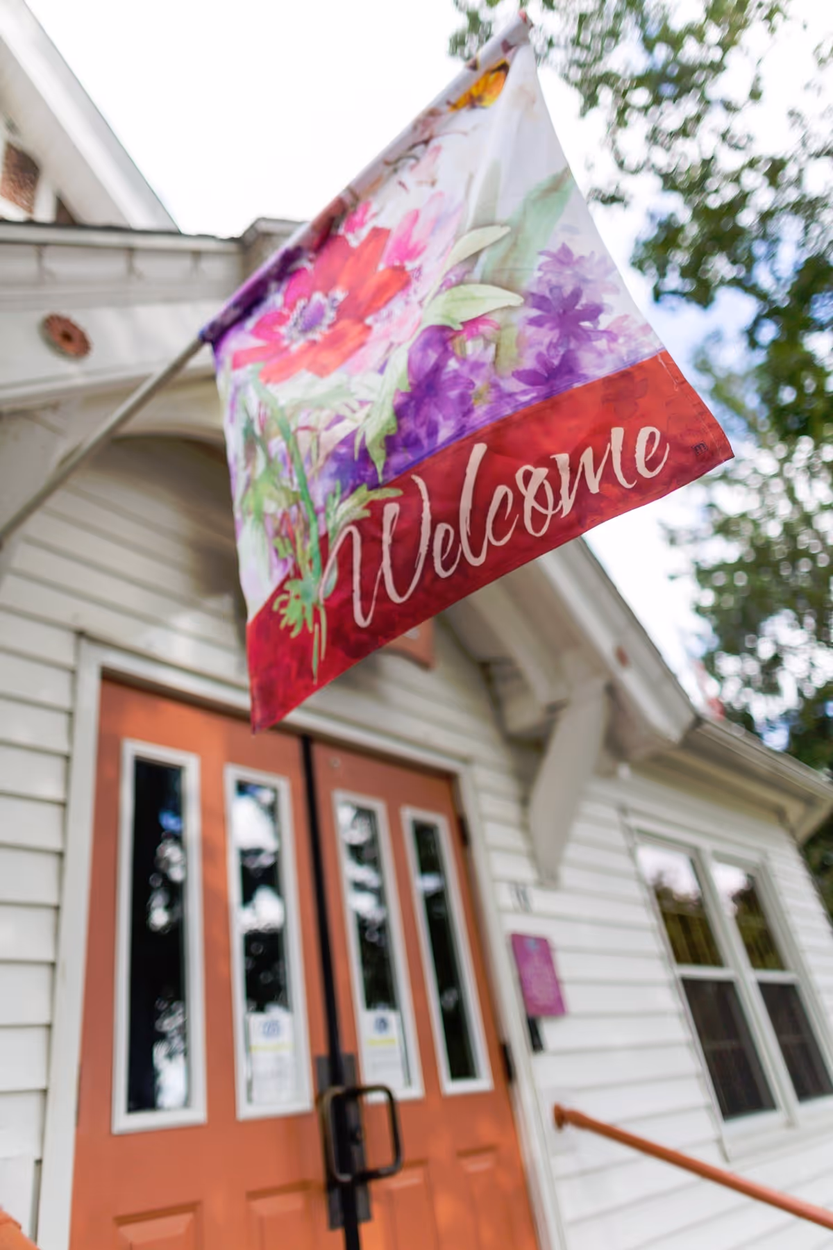 A colorful floral "Welcome" flag hangs above the front entrance of a white-sided building with double red doors.