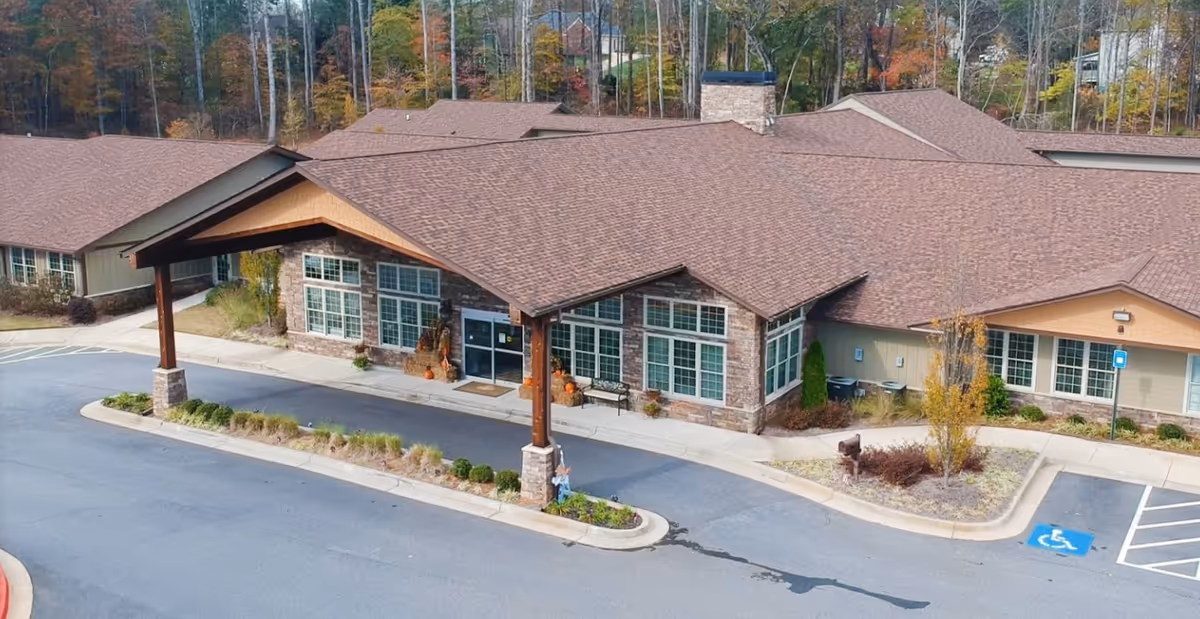 Front entrance of a single-story senior living building with a covered porte-cochere, large windows, parking spaces, and landscaped beds.