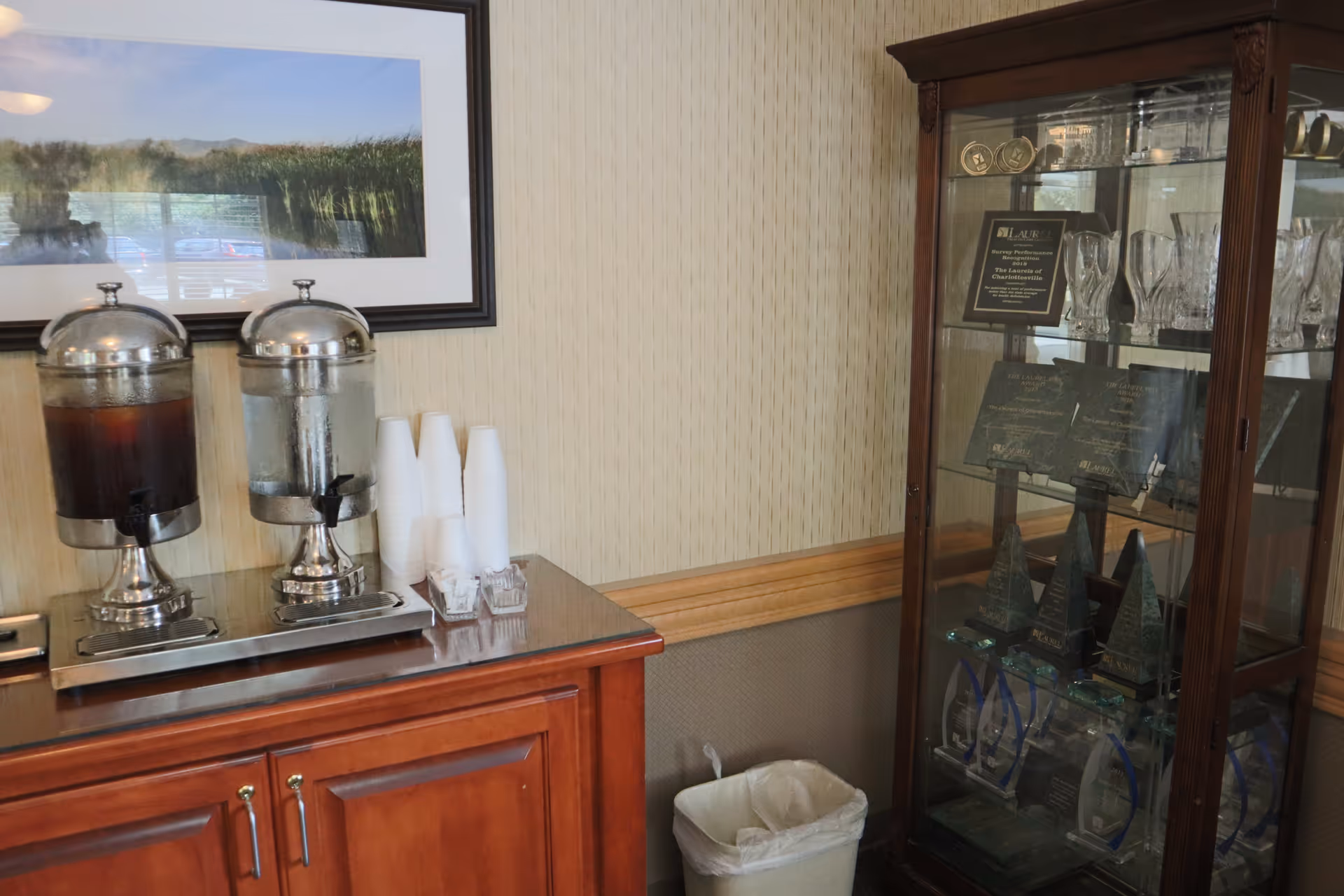 A refreshment station with two beverage dispensers, one filled with iced tea and the other with water, next to a stack of white disposable cups. To the right, there is a glass display cabinet containing various trophies and awards. A framed landscape photograph hangs on the wall above the beverage dispensers.
