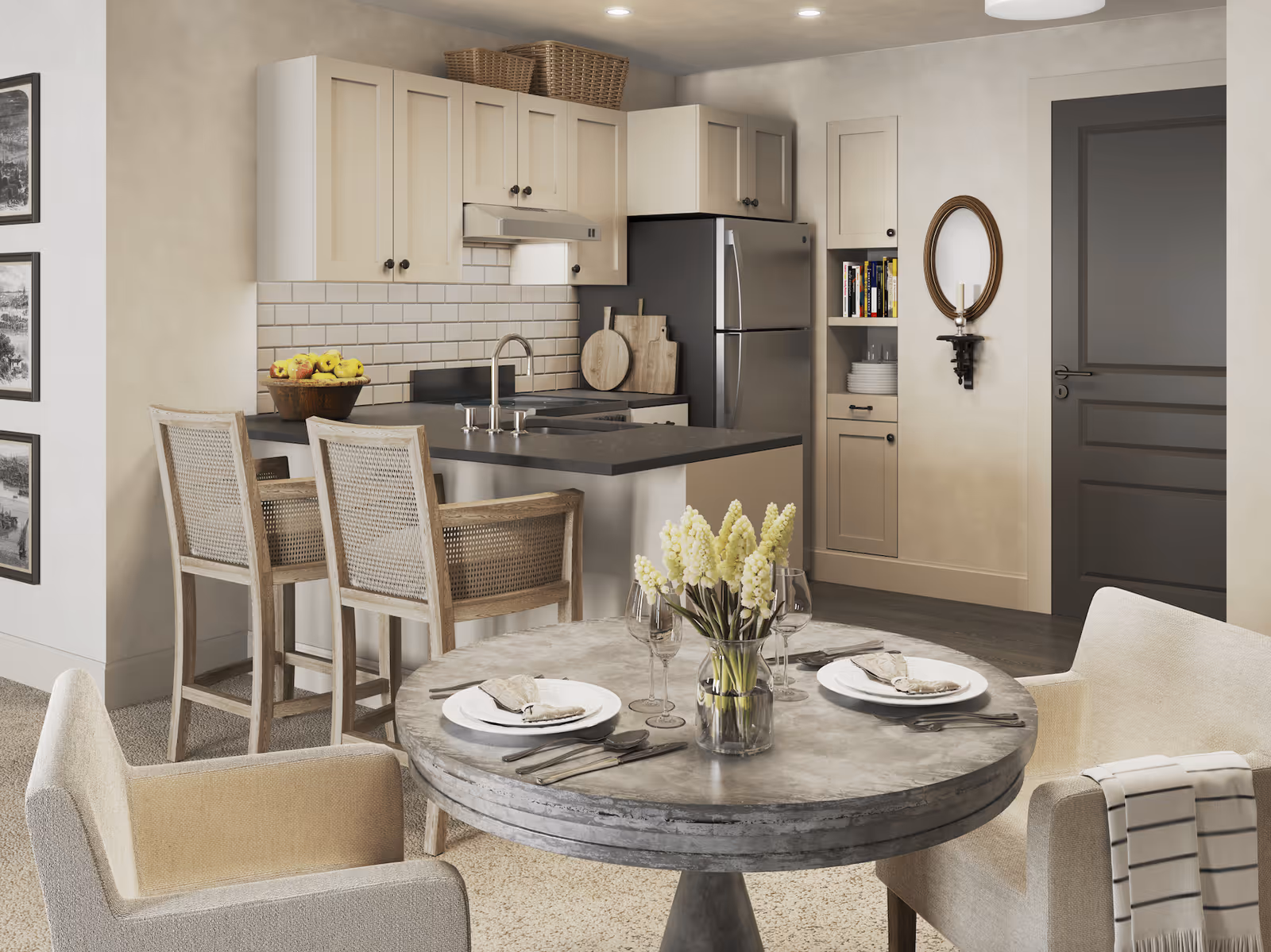 A modern kitchen and dining area featuring a round dining table set for two with plates, napkins, cutlery, wine glasses, and a vase of white flowers. The kitchen has light-colored cabinets, a stainless steel refrigerator, a black countertop with a sink, two wooden bar stools, and decorative baskets on top of the cabinets. A dark door and a wall-mounted oval mirror with a candle holder are visible in the background.