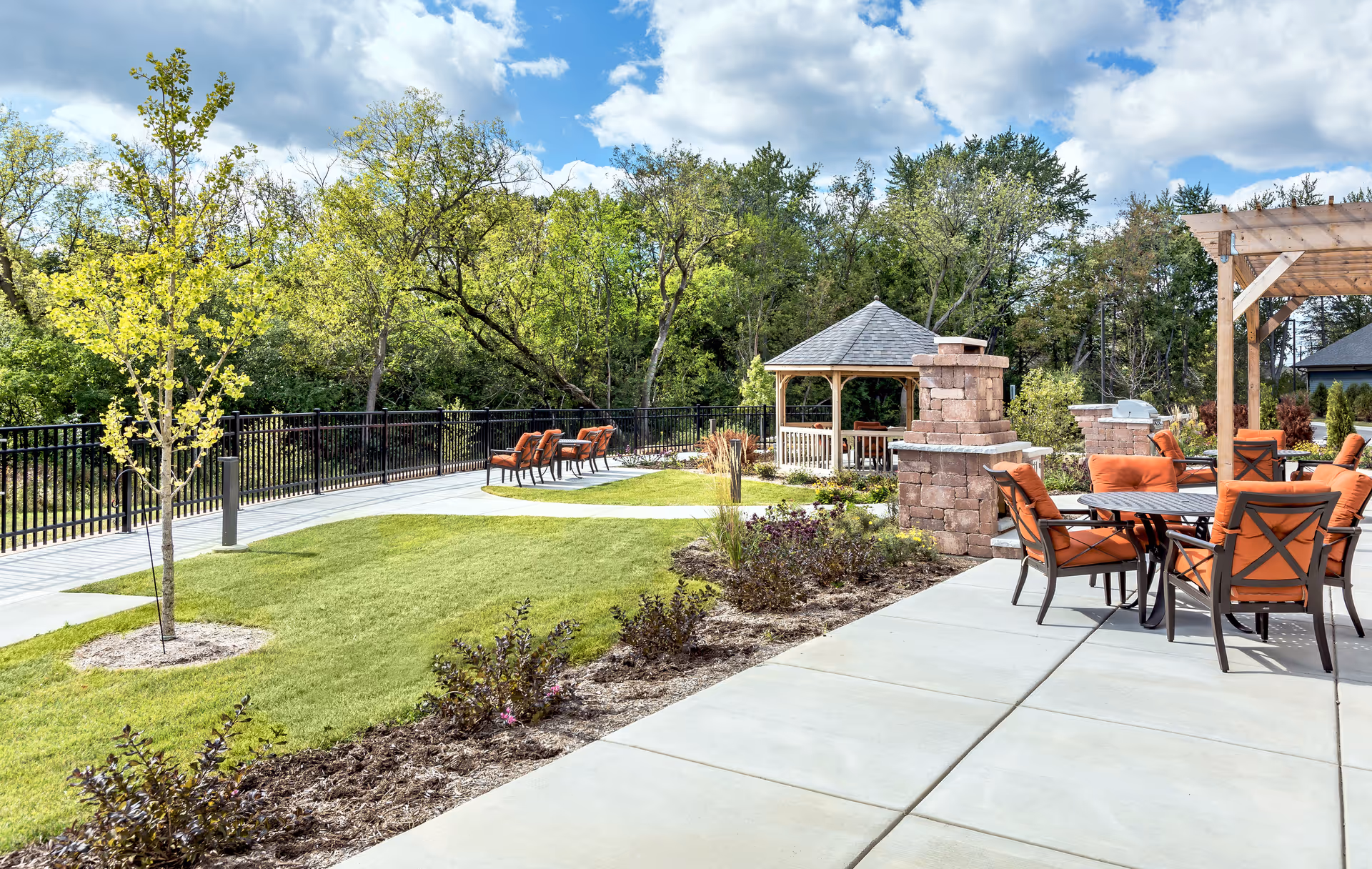 Outdoor patio area with orange cushioned chairs around tables, a stone fireplace, a wooden pergola, a gazebo in the background, surrounded by green grass, trees, and a clear blue sky with some clouds.