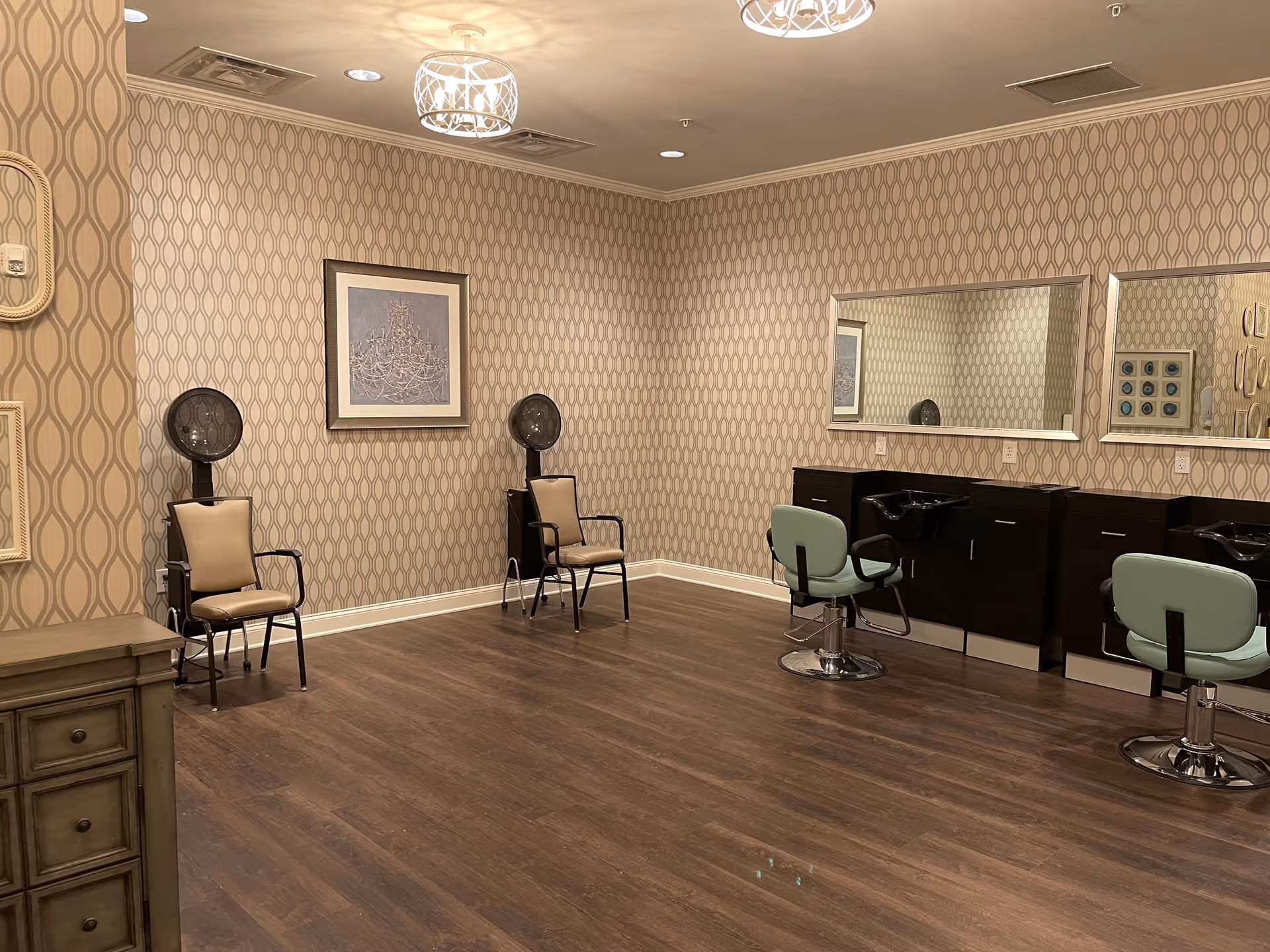 Interior of a salon room with two hair drying chairs against a patterned wallpapered wall, two salon styling chairs in front of large mirrors, wooden flooring, and ceiling lights.