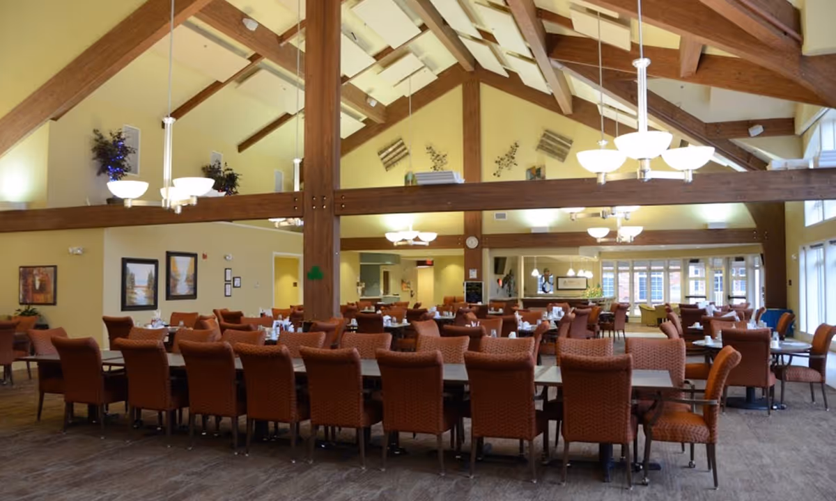A spacious dining room with high vaulted ceilings featuring exposed wooden beams and multiple hanging light fixtures. The room is furnished with several long tables surrounded by cushioned chairs upholstered in a reddish-brown fabric. The walls are painted a soft yellow and decorated with framed artwork and plants. Large windows on the right side allow natural light to fill the space.