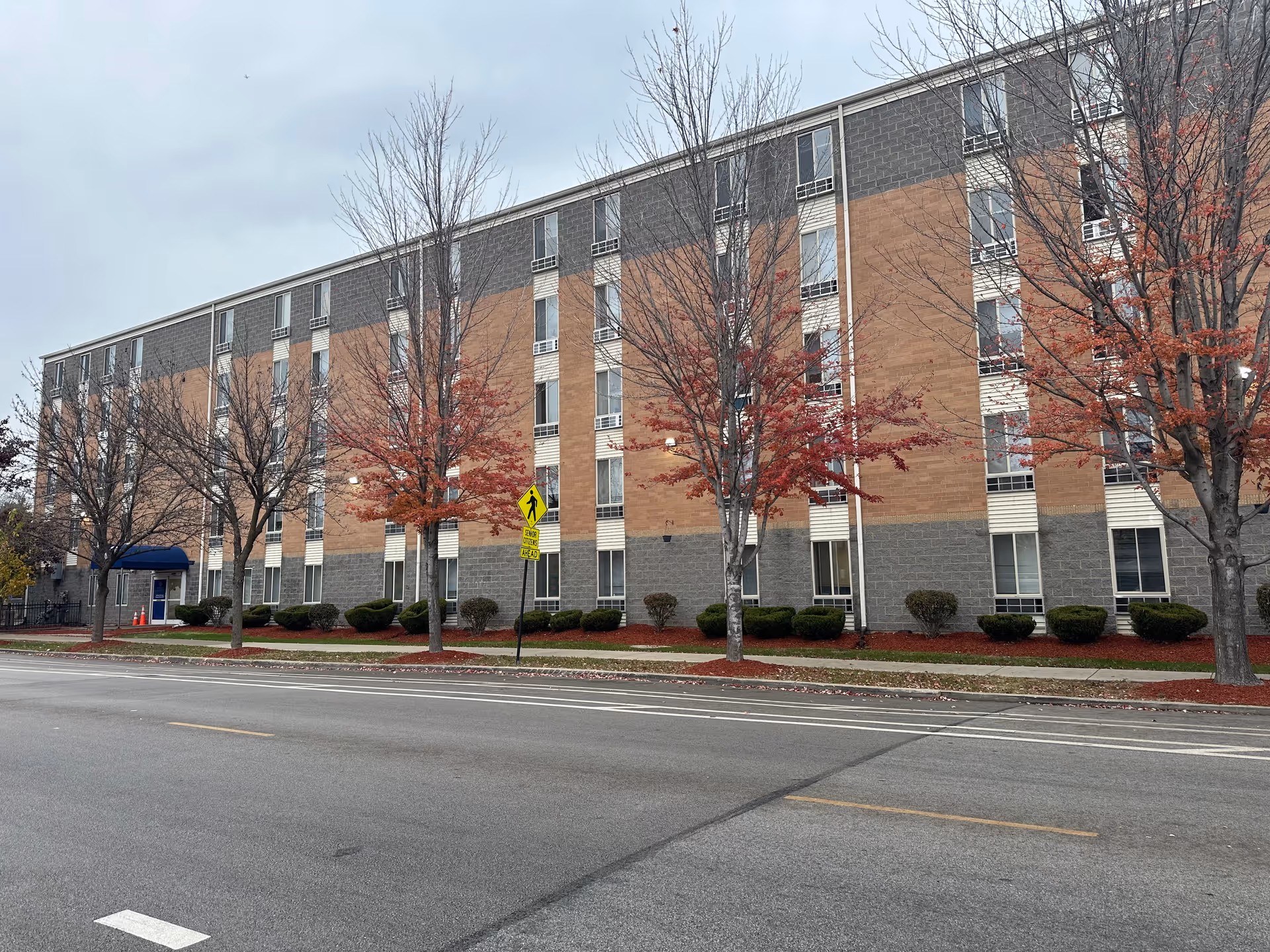 Exterior view of a multi-story supportive living facility building with a brick and gray facade, several windows, leafless trees with some red leaves, and a pedestrian crossing sign near the sidewalk on a cloudy day.