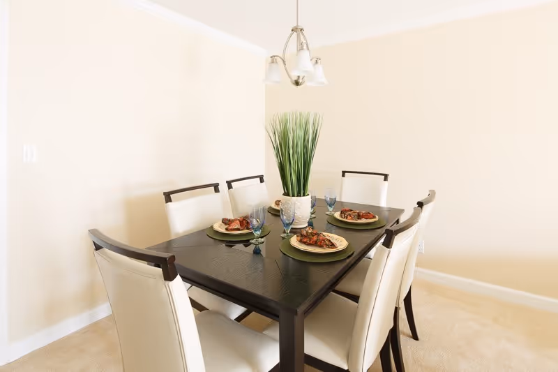 Dining room with a dark wood table set with four place settings, six white upholstered chairs, a potted centerpiece and an overhead chandelier.