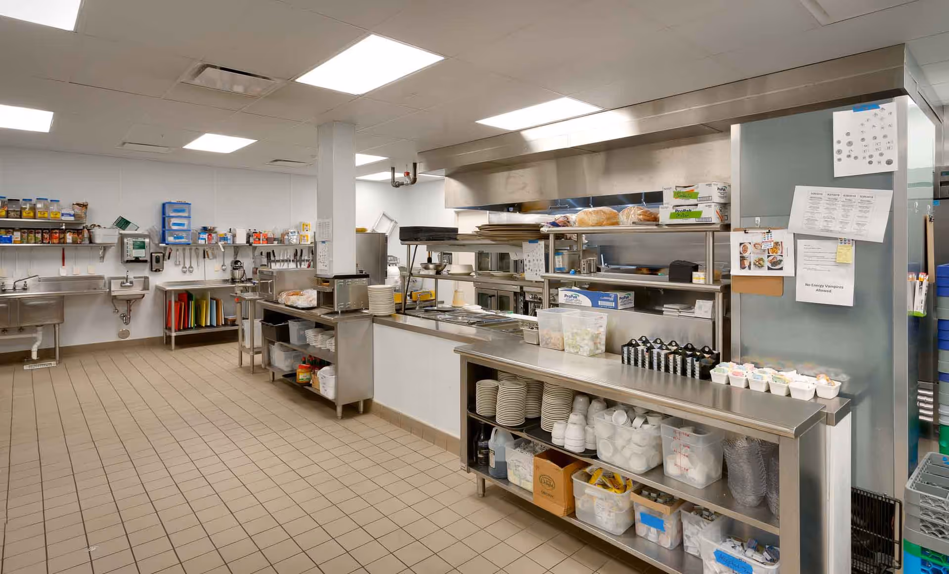 Commercial stainless-steel kitchen with prep counters, shelving stocked with dishes and supplies, sinks, and cooking equipment.