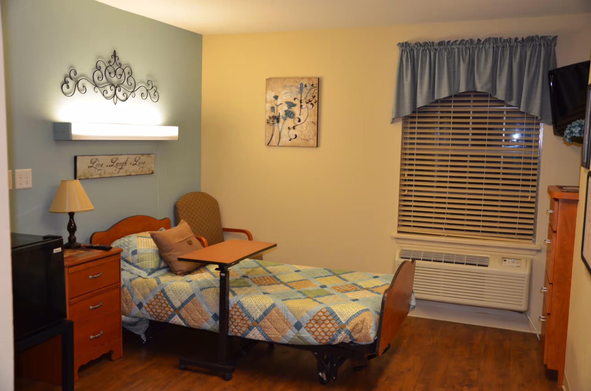 A nursing facility bedroom with a single bed covered in a patchwork quilt. Next to the bed is a wooden nightstand with a lamp and a small black refrigerator. There is a cushioned chair with a small overbed table in front of it. The walls are painted in two tones, light blue and beige, with decorative wall art and a sign that reads 'Live Laugh Love'. A window with blinds and a blue valance is above an air conditioning unit. A flat-screen TV is mounted on the wall to the right.