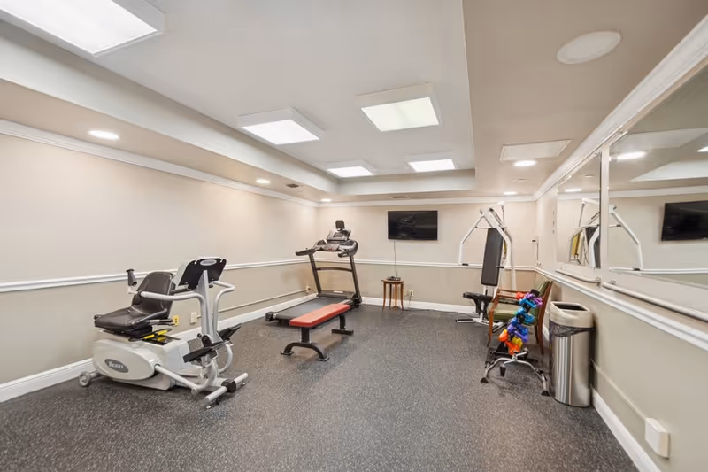 A small fitness room with exercise equipment including a recumbent bike, treadmill, weight bench, multi-function weight machine, and a rack of colorful dumbbells. The room has beige walls, a large mirror on one side, recessed lighting, and a wall-mounted TV.