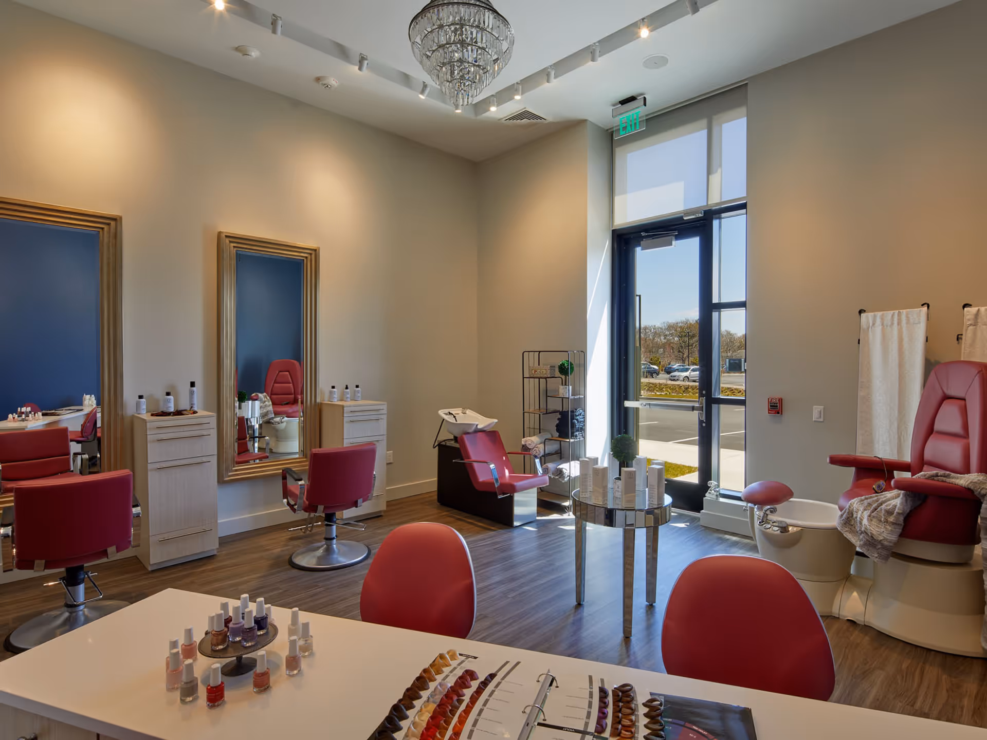 A bright and modern salon area inside a senior living facility with red salon chairs, large mirrors, a pedicure station with a red chair and foot bath, a table displaying nail polish bottles and color samples, and a glass door letting in natural light.