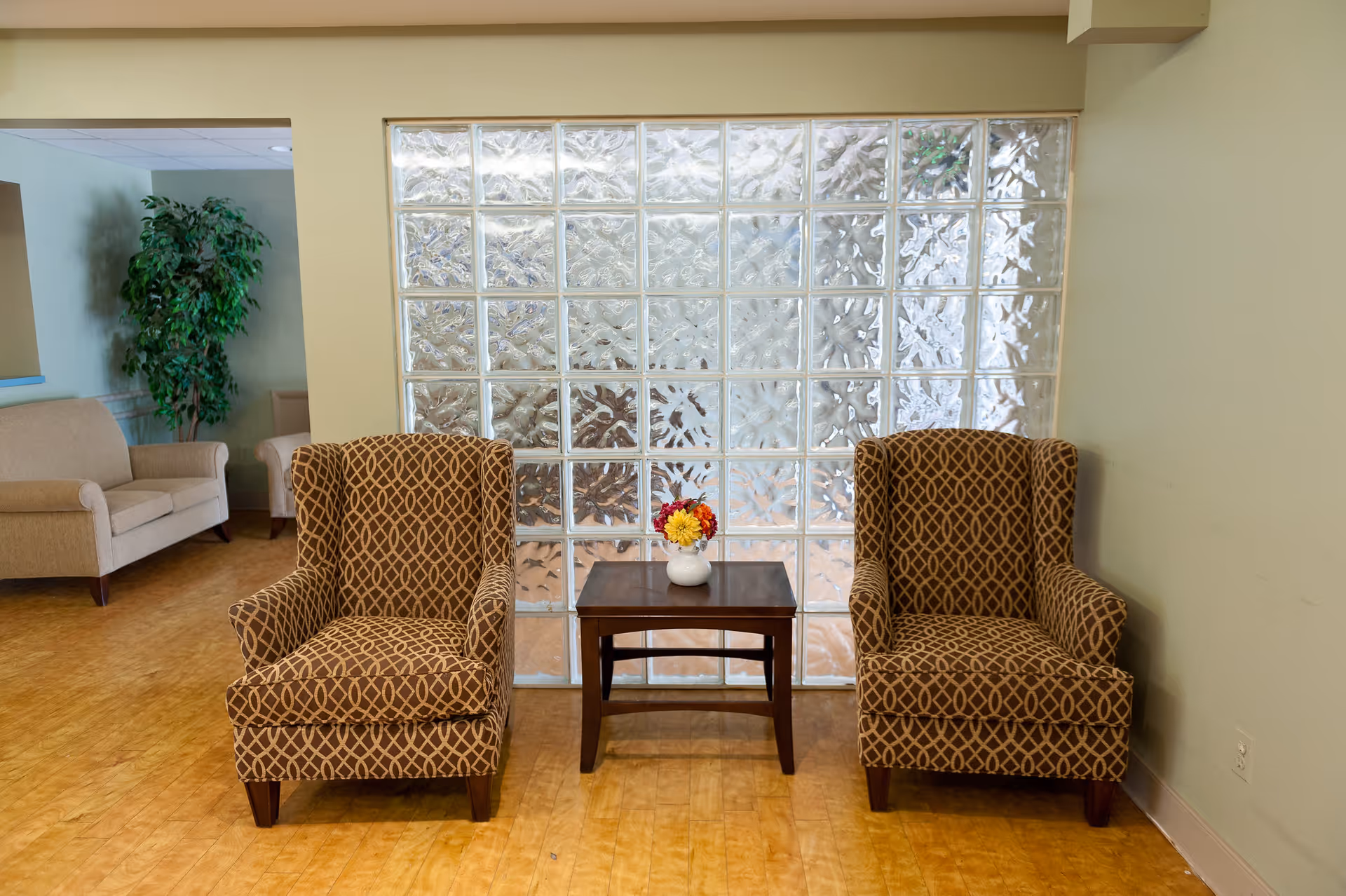 Two patterned armchairs with a small wooden table between them holding a small vase with colorful flowers, set against a wall made of glass blocks. In the background, there is a beige sofa and a green potted plant in a room with light-colored walls and wooden flooring.