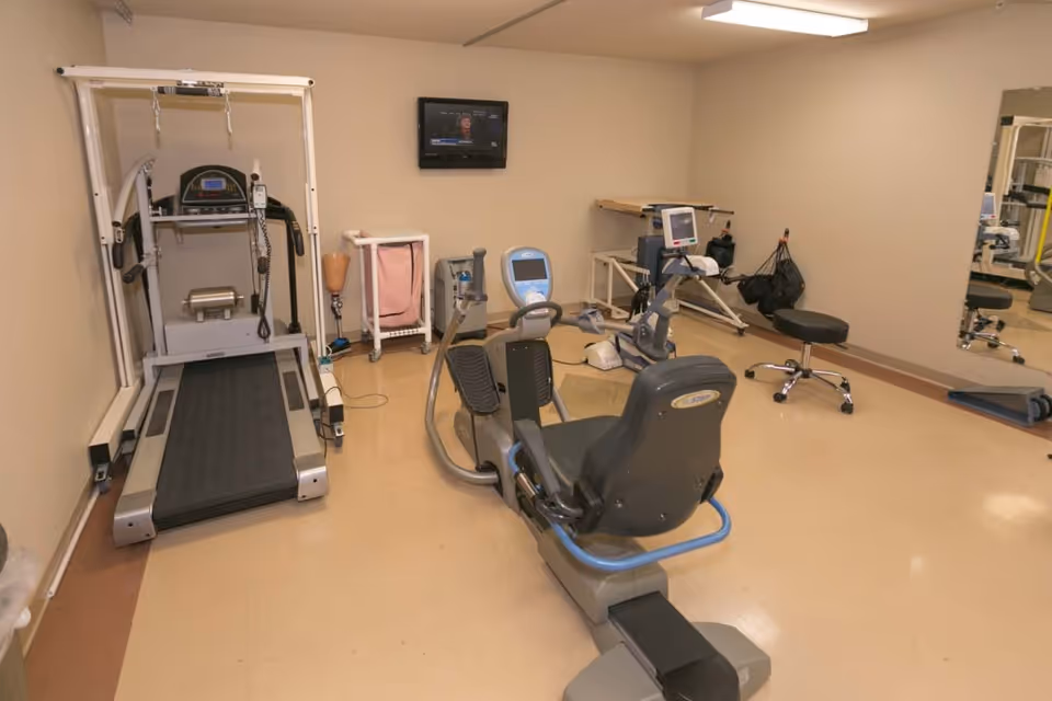 A small exercise room with a treadmill, a recumbent exercise bike, a stationary bike, a stool, and various medical and exercise equipment. A small television is mounted on the beige wall, and a large mirror is on the right side of the room.