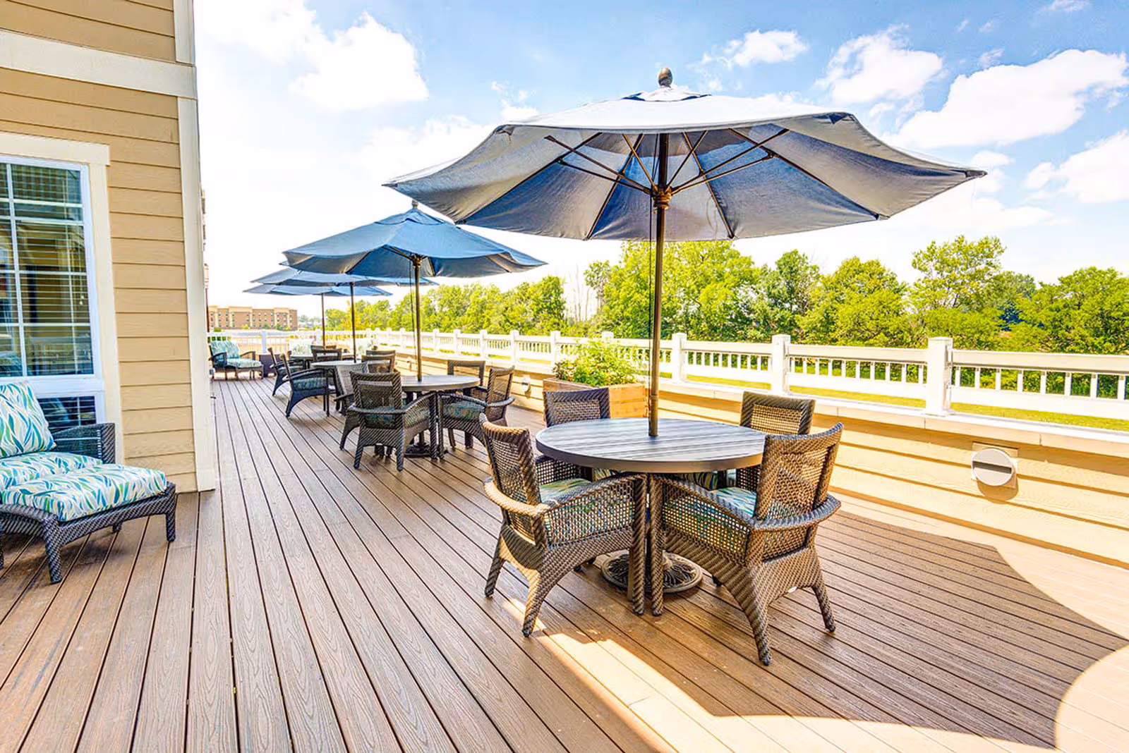 Sunny outdoor deck with round tables, wicker chairs and large blue umbrellas overlooking trees.