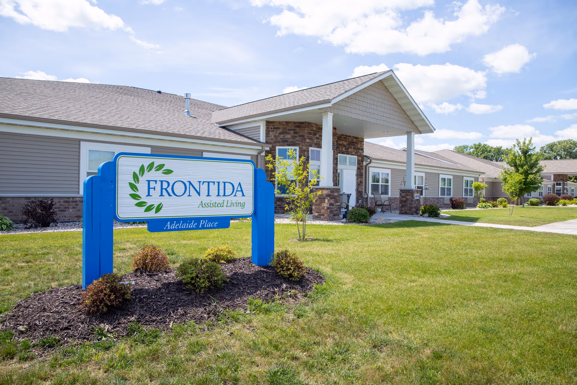 Exterior view of Frontida Assisted Living: Adelaide Place building with a blue and white sign in front, surrounded by grass, small bushes, and a clear sky with some clouds.