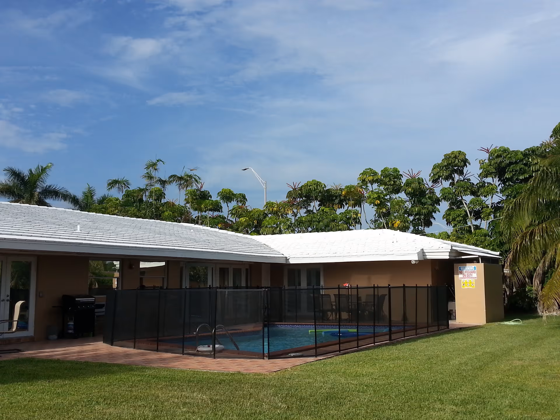 Outdoor view of a single-story building with a white roof and a fenced swimming pool in the backyard. The pool area is surrounded by a black safety fence, and there is green grass in the foreground. Trees and a clear blue sky are visible in the background.