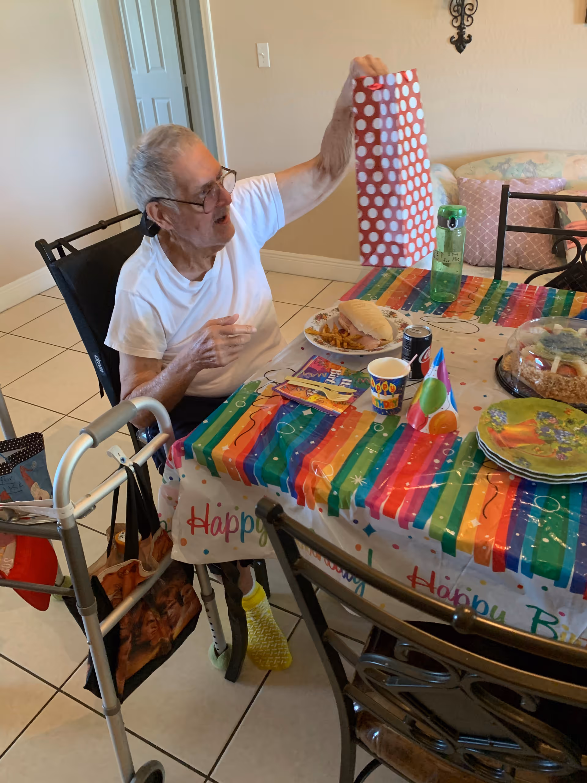 An elderly person sitting in a wheelchair at a dining table decorated with a colorful birthday-themed tablecloth and party items, holding up a red and white polka dot gift bag. The table has a sandwich, fries, a soda can, a water bottle, a birthday cup, party hats, and a cake. A walker with bags hanging on it is next to the person.