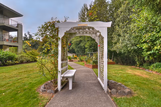 A paved garden pathway passing under a white wooden arbor with lattice sides and a bench attached. The pathway leads to raised garden beds surrounded by green grass, shrubs, and trees. A multi-story building with balconies is visible on the left side under a cloudy sky.