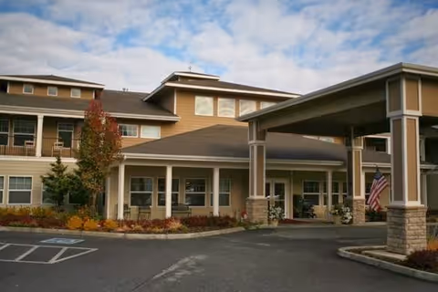 Exterior front entrance of a multi-story senior living building with a covered porte-cochere, landscaping, and an American flag.