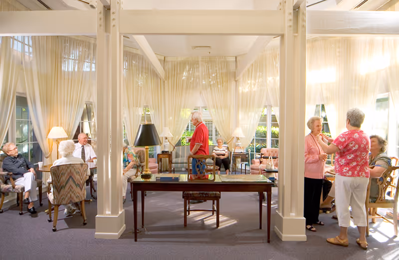 A bright and spacious common area in a senior living facility with large windows and sheer curtains. Several elderly individuals are seated and engaged in conversation, while two women stand and talk near the right side. The room is furnished with comfortable chairs, side tables with lamps, and a central wooden table with a lamp and books.
