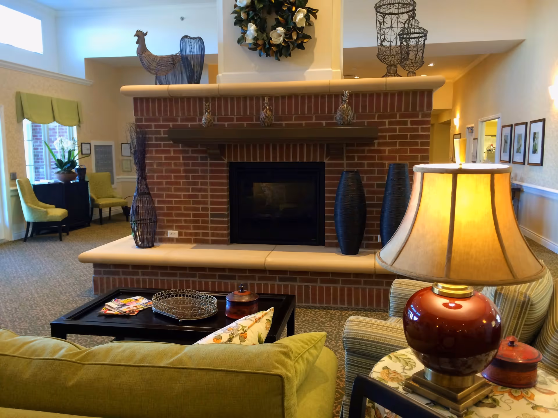 A cozy living room area with a brick fireplace as the focal point. The fireplace mantel is decorated with small pineapples and wire sculptures. In front of the fireplace is a coffee table with magazines and decorative items. There is a green couch with a floral pillow, a side table with a red lamp, and two green chairs near a window with a green valance. The room has warm lighting and framed pictures on the wall.