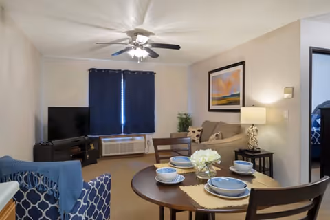 A cozy living room and dining area in a senior living facility. The room features a round dining table set with blue and white dishes and a small flower arrangement. There is a blue patterned armchair, a beige sofa with decorative pillows, a side table with a lamp, a flat-screen TV on a stand, and dark curtains covering a window. A ceiling fan with lights is mounted on the ceiling.
