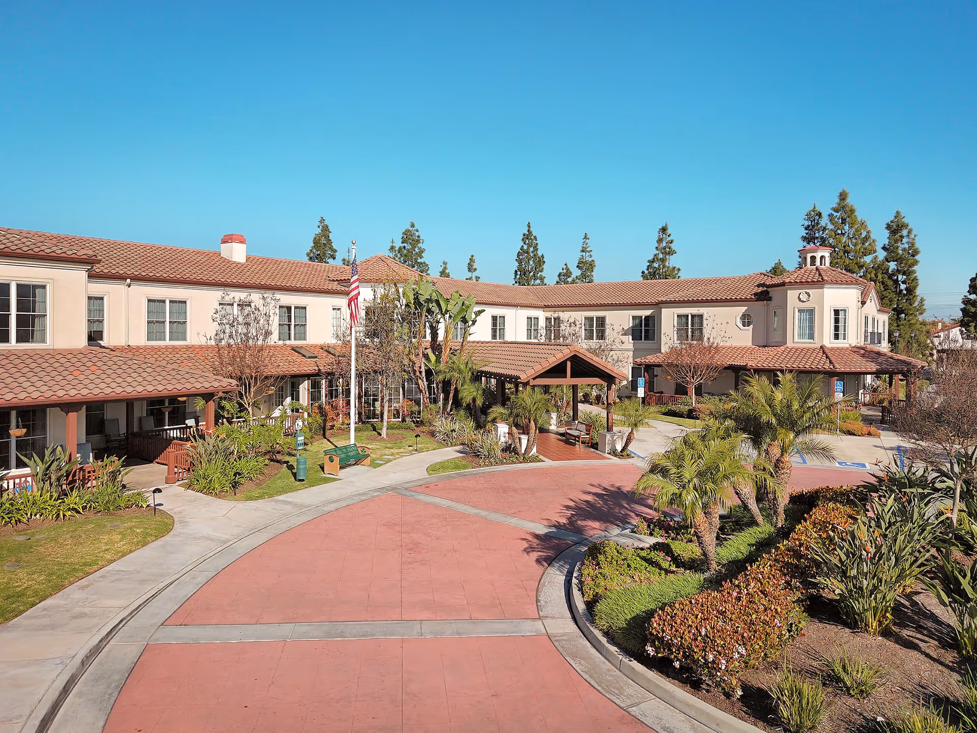 Exterior view of a senior living facility named Ivy Park at La Palma, featuring a two-story building with a red tile roof, beige walls, and multiple windows. The entrance area has a covered walkway, an American flag on a flagpole, landscaped greenery, palm trees, and a curved driveway with a red pavement under a clear blue sky.