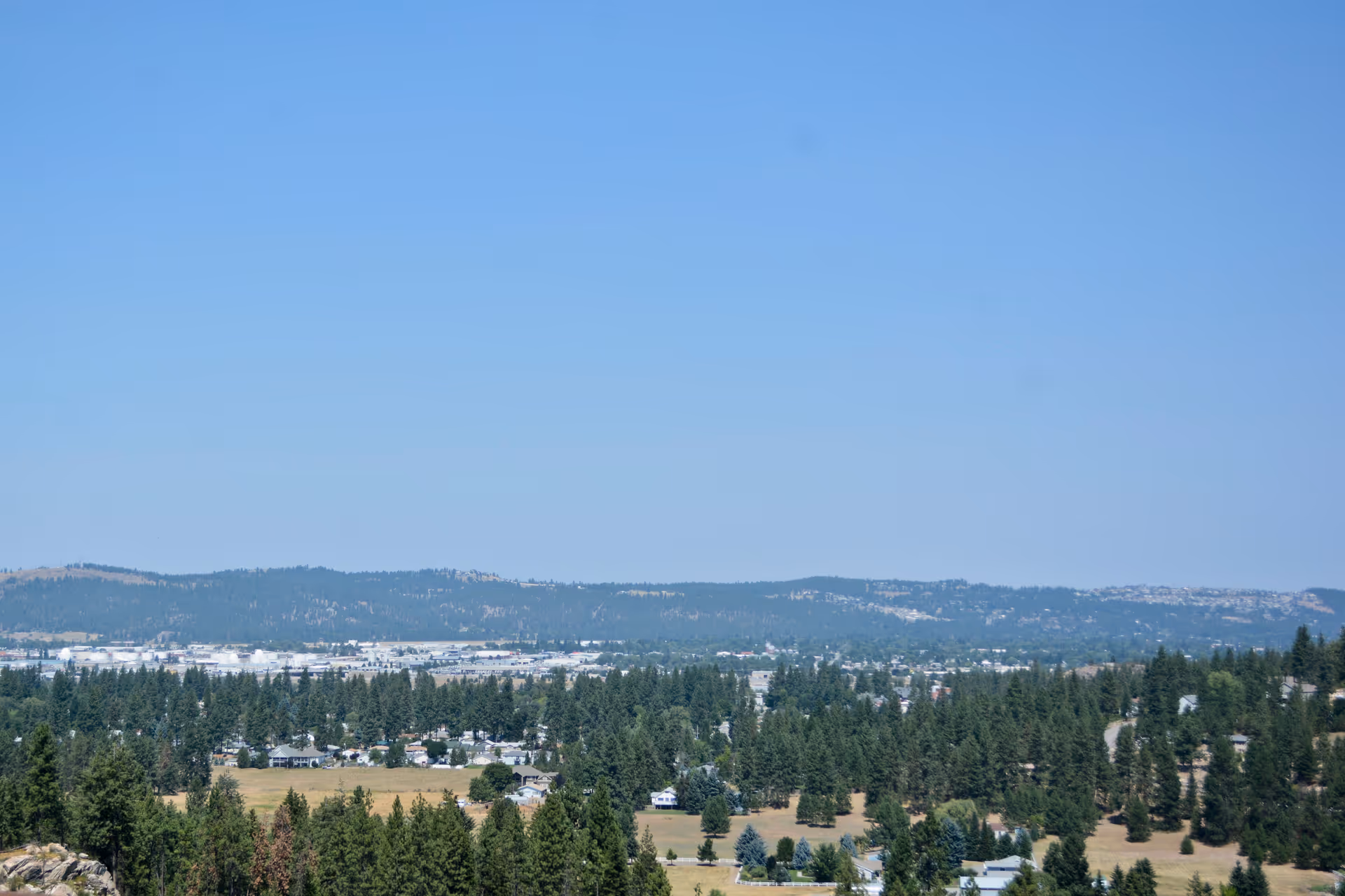 A wide landscape view showing a forested area with many trees, scattered houses, and a distant mountain range under a clear blue sky.