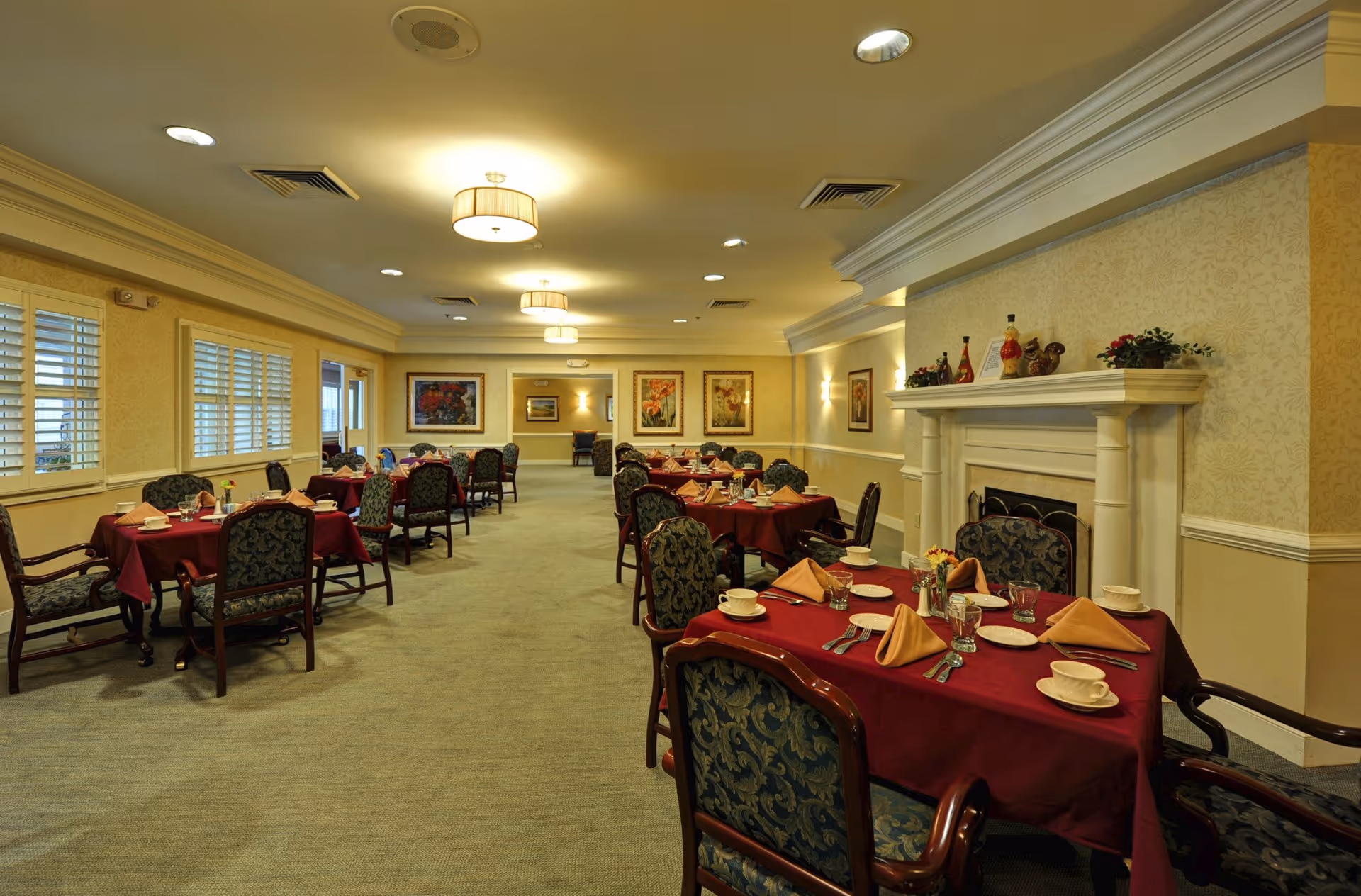 Formal dining room with multiple set tables covered in red tablecloths, upholstered chairs, a fireplace, and framed artwork on the walls.