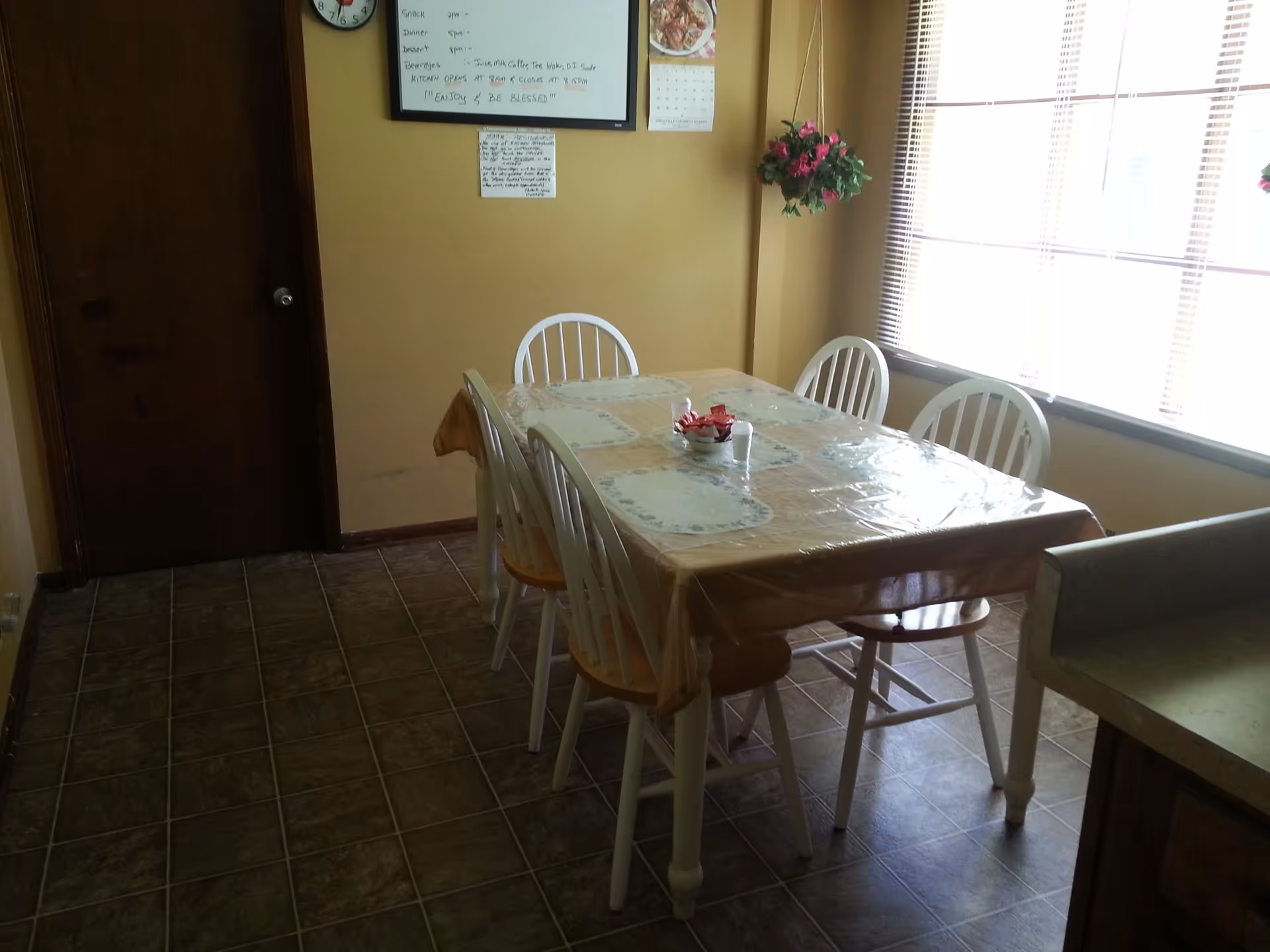 Small dining area with a rectangular table covered in plastic, six white chairs, a large window and a door.