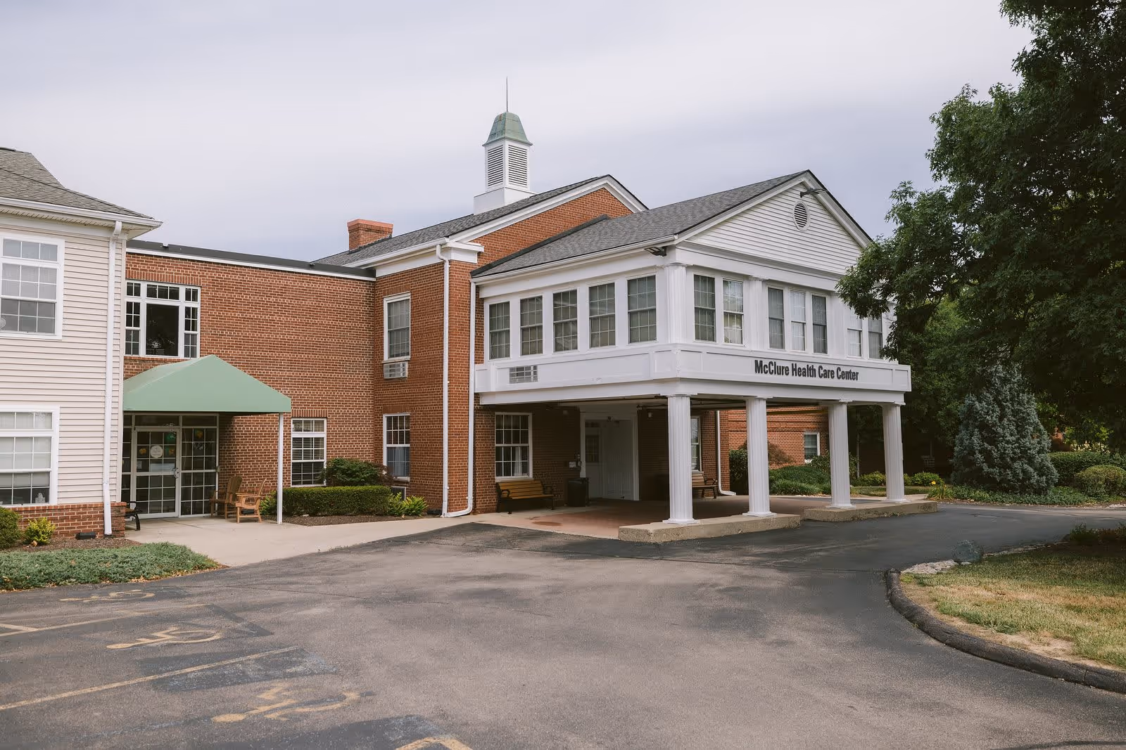 Exterior view of the McClure Health Care Center building at Ohio Living Mount Pleasant, featuring a brick facade with white trim, a covered entrance supported by white columns, and surrounding greenery including trees and shrubs.