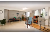 Bright physical therapy/exercise room with parallel bars, chairs, therapy equipment, and plants by large windows.
