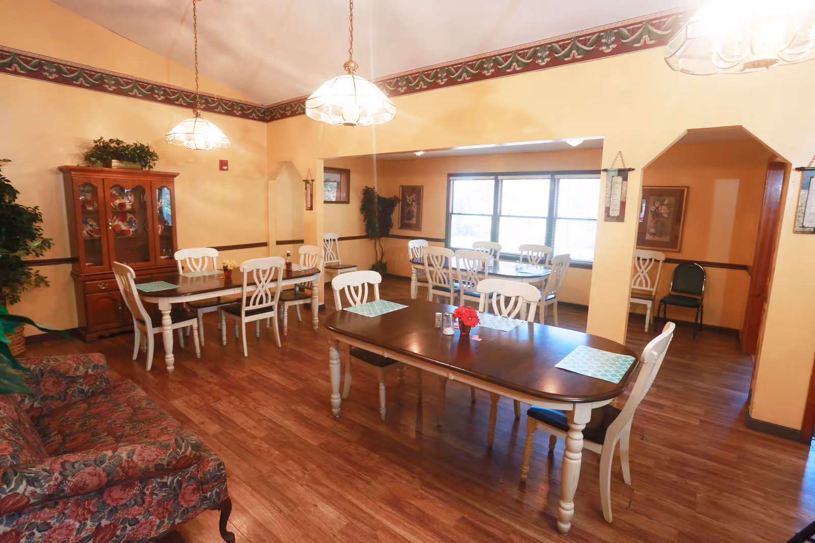 Communal dining room with several wooden tables and white chairs, pendant lights, a china cabinet and windows.
