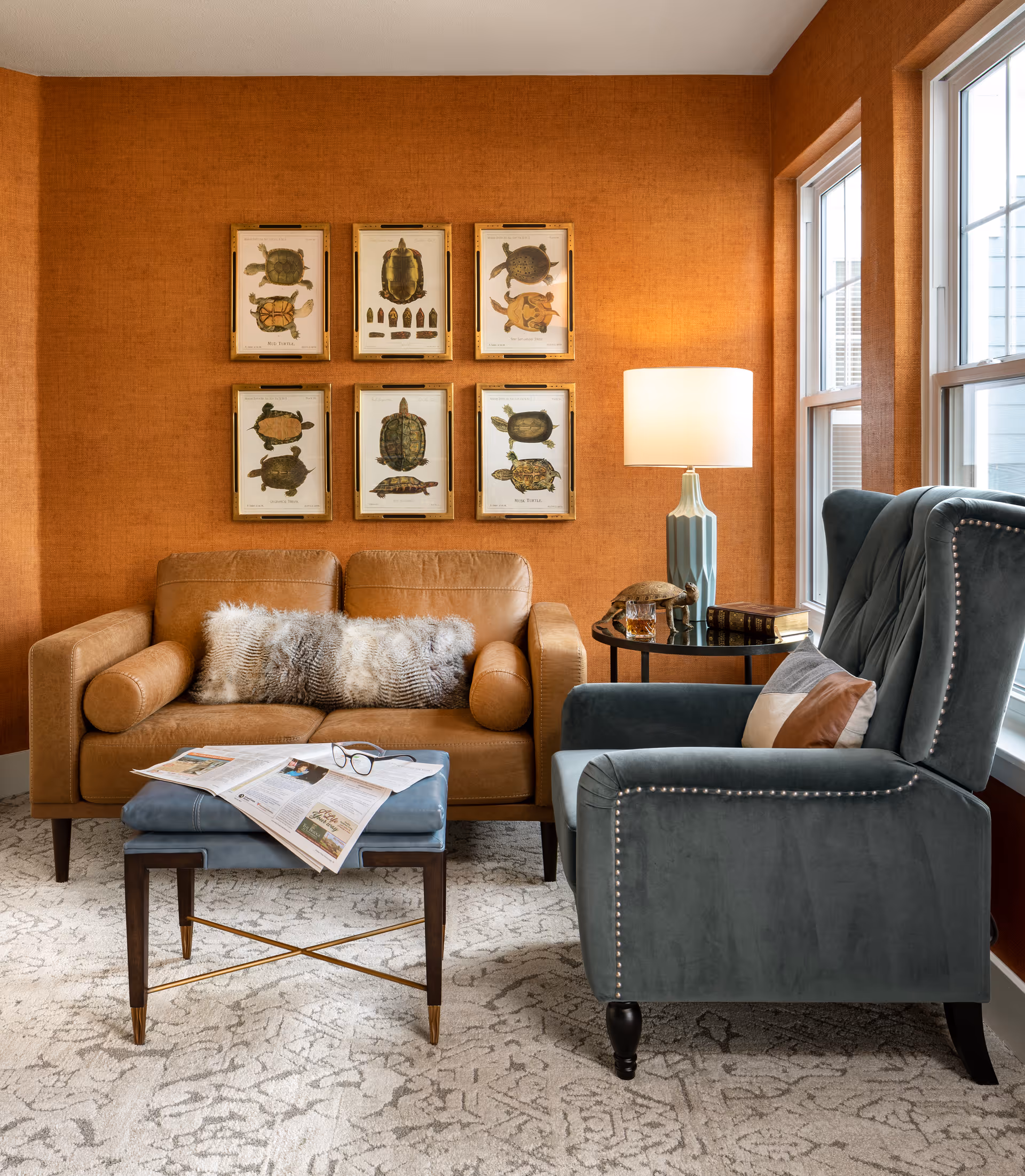 A cozy living room area with an orange textured wall featuring six framed turtle illustrations. There is a tan leather loveseat with a fluffy pillow, a blue upholstered armchair with a decorative pillow, and a small blue ottoman with a newspaper and glasses on it. A round side table with a blue lamp, books, and a small turtle figurine is placed between the loveseat and armchair. Large windows allow natural light to brighten the space.