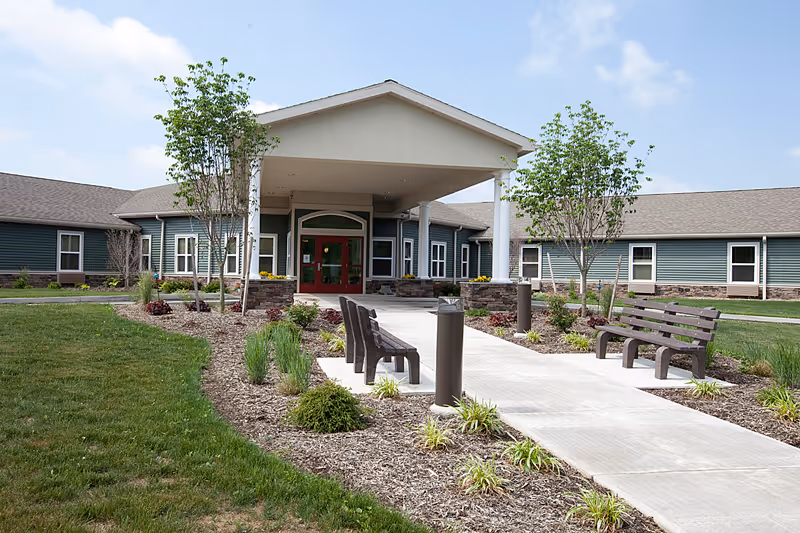 Exterior view of the entrance to Wolf Run Village Senior Living Community, showing a covered walkway with benches on either side, landscaped garden beds with small trees and plants, and a clear blue sky.