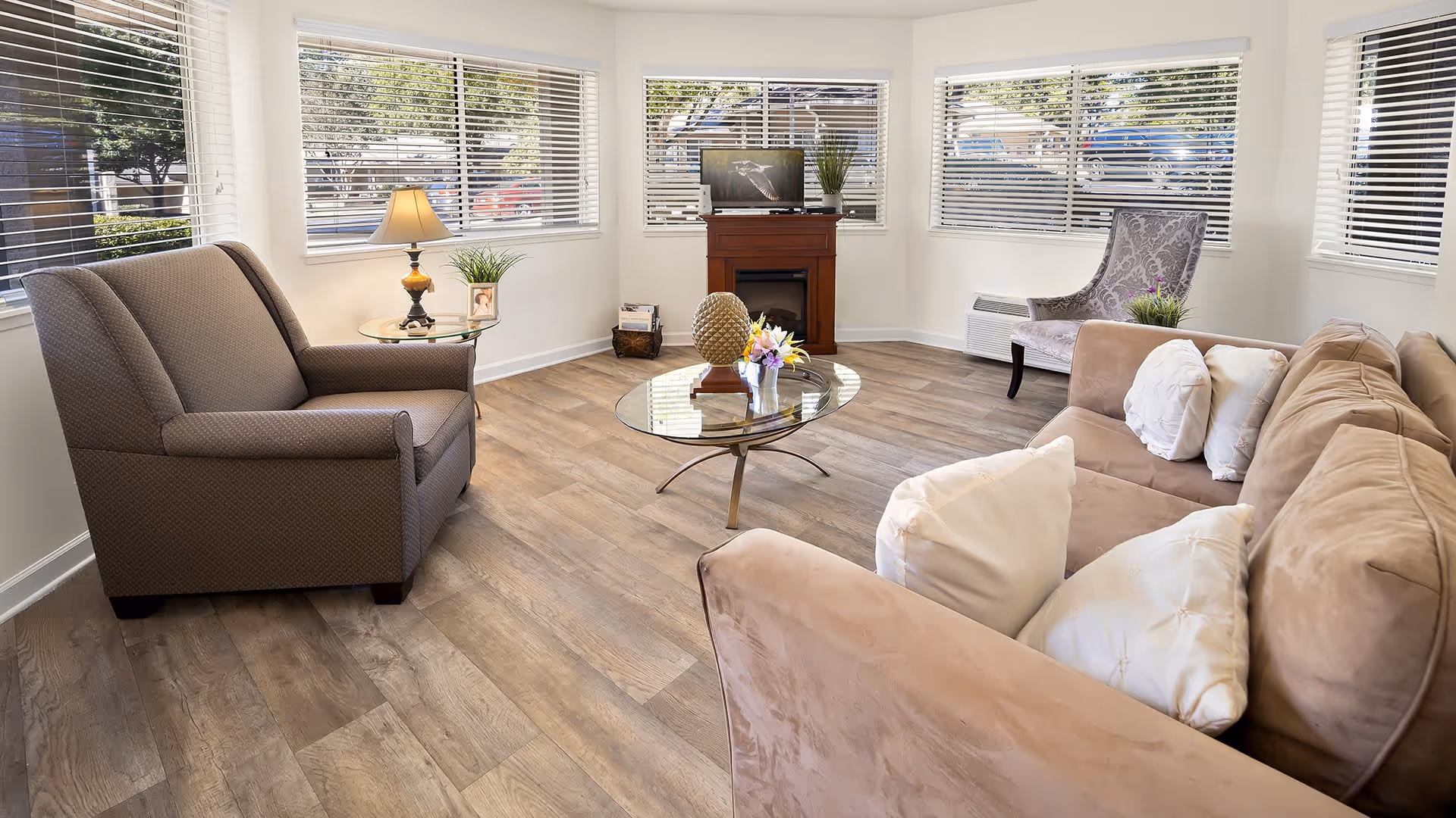 A bright and cozy living room with large windows covered by white blinds. The room features a beige sofa with white pillows, a brown armchair, a patterned accent chair, a glass coffee table with decorative items, a side table with a lamp and plant, and a small wooden TV stand with a flat-screen TV and a plant on top. The floor is wood with a natural finish.