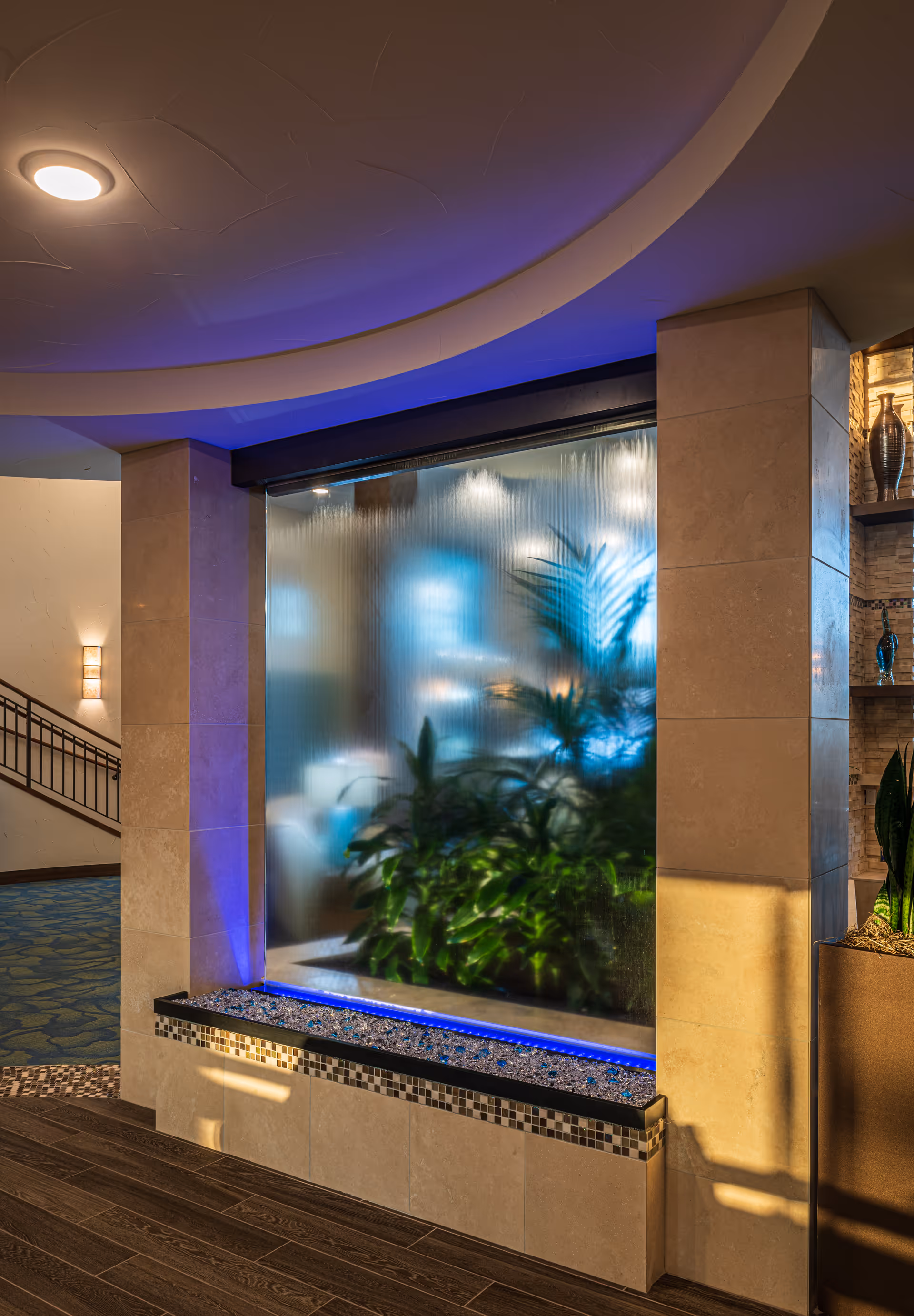 Indoor water feature with a vertical glass panel and plants behind it, illuminated with blue lighting. The surrounding area includes tiled walls, a wooden floor, and decorative vases on shelves to the right. A staircase with a railing is visible in the background to the left.