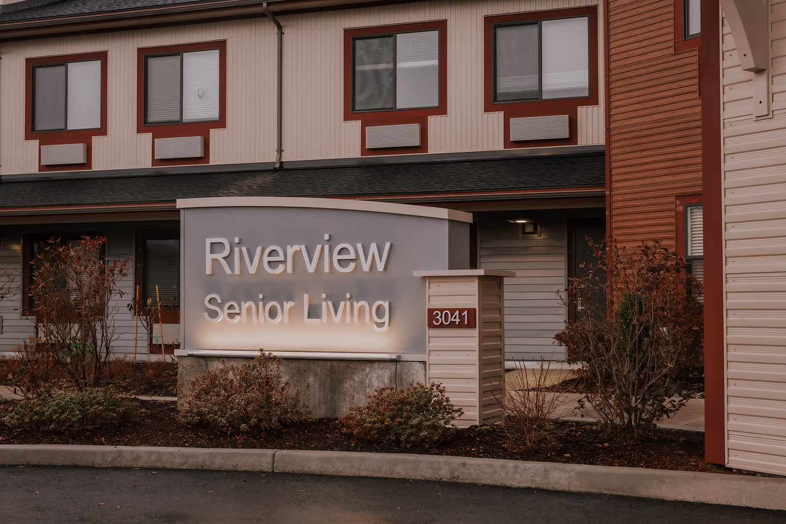 A lit sign reading 'Riverview Senior Living' with the address '3041' sits in front of a two-story senior living building.