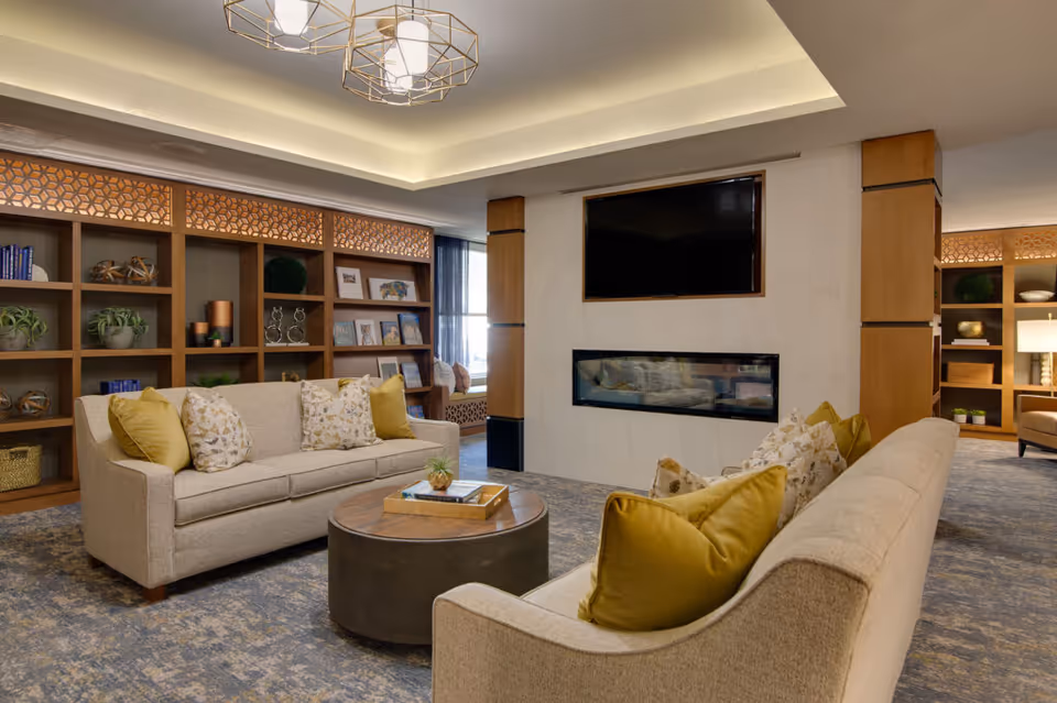 A cozy living room area with two beige sofas adorned with yellow and patterned throw pillows facing a modern fireplace and a wall-mounted flat-screen TV. Behind the sofas, there are wooden built-in shelves decorated with plants, books, and decorative items. The room features a soft blue and beige carpet, warm lighting, and a contemporary ceiling light fixture.