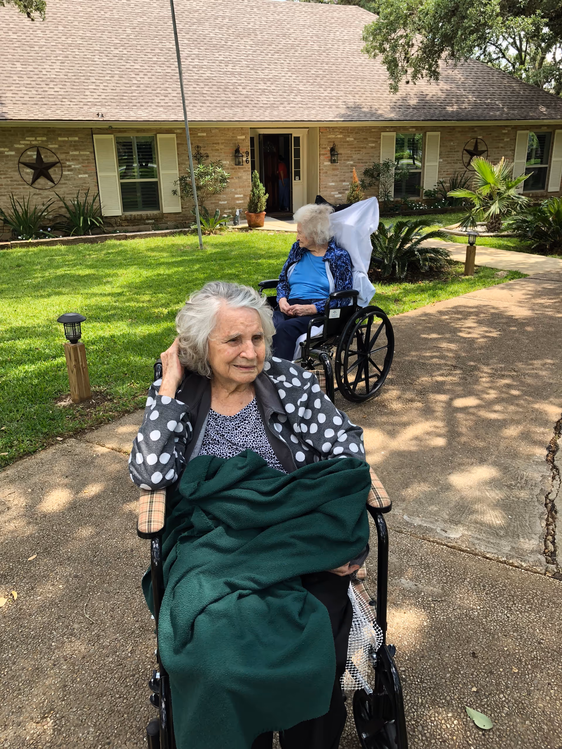 Two elderly women sitting in wheelchairs outside on a paved pathway in front of a single-story brick house with a well-maintained lawn and plants. One woman is covered with a green blanket and wearing a polka dot jacket, while the other woman is wearing a blue top and a patterned jacket. The scene is shaded by trees.
