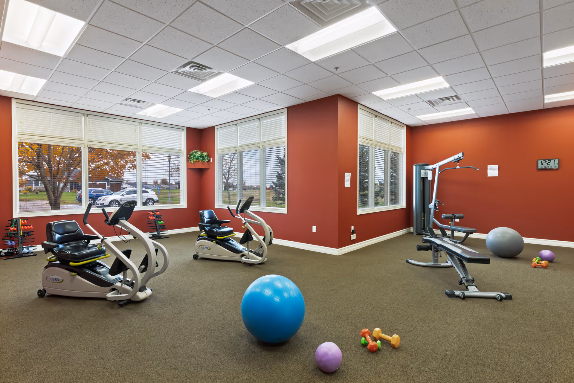 A senior living facility exercise room with two recumbent stationary bikes, a weight bench with attached pulleys, various small dumbbells, and exercise balls on a carpeted floor. The room has large windows with white blinds, red walls, and ceiling lights.