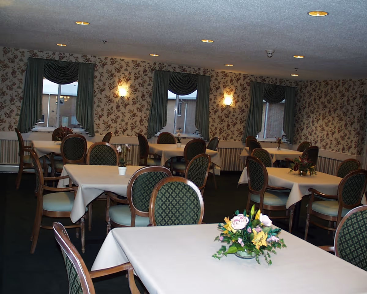 Interior view of a dining room with multiple tables covered with white tablecloths and decorated with floral centerpieces. The room has floral wallpaper, green curtains on the windows, and green cushioned chairs with wooden frames arranged around the tables. Ceiling lights illuminate the space.