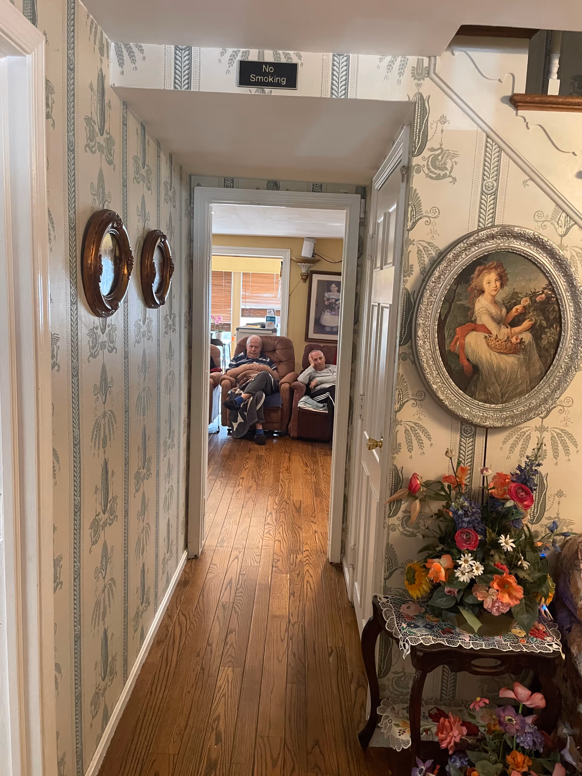 A hallway with patterned wallpaper and wooden floor leading to a living room area where two elderly men are seated in recliner chairs. On the right wall of the hallway, there is a round framed painting of a young girl holding flowers and a basket, above a small table with a floral arrangement. Two small round mirrors hang on the left wall. A 'No Smoking' sign is visible above the doorway.