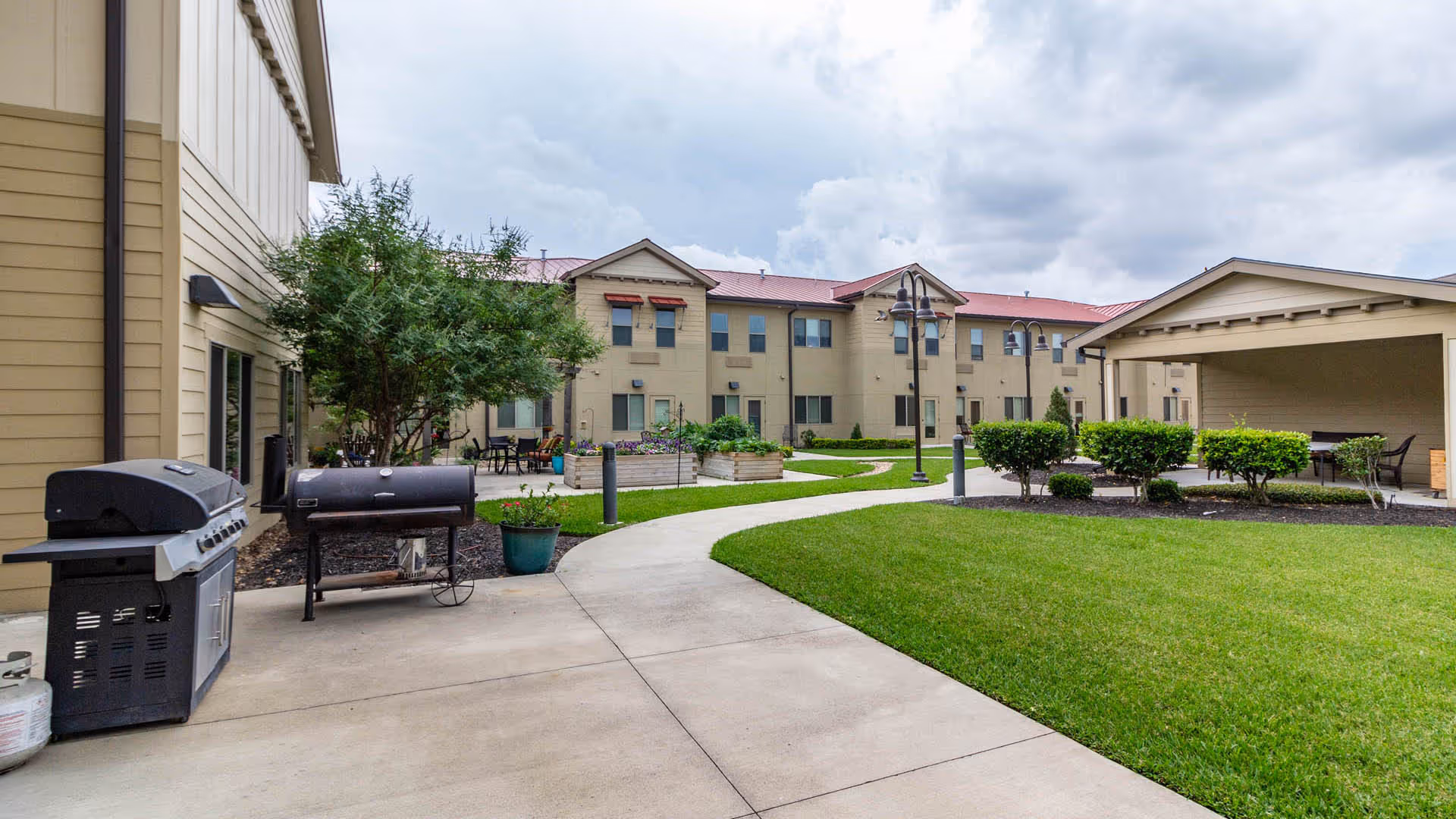 Outdoor courtyard area of a senior living facility with a concrete walkway winding through green grass and landscaped bushes. There are two barbecue grills on the left side near the building, raised garden beds with flowers, patio tables and chairs, and a covered pavilion on the right. The building is two stories with beige siding and red roof, under a cloudy sky.
