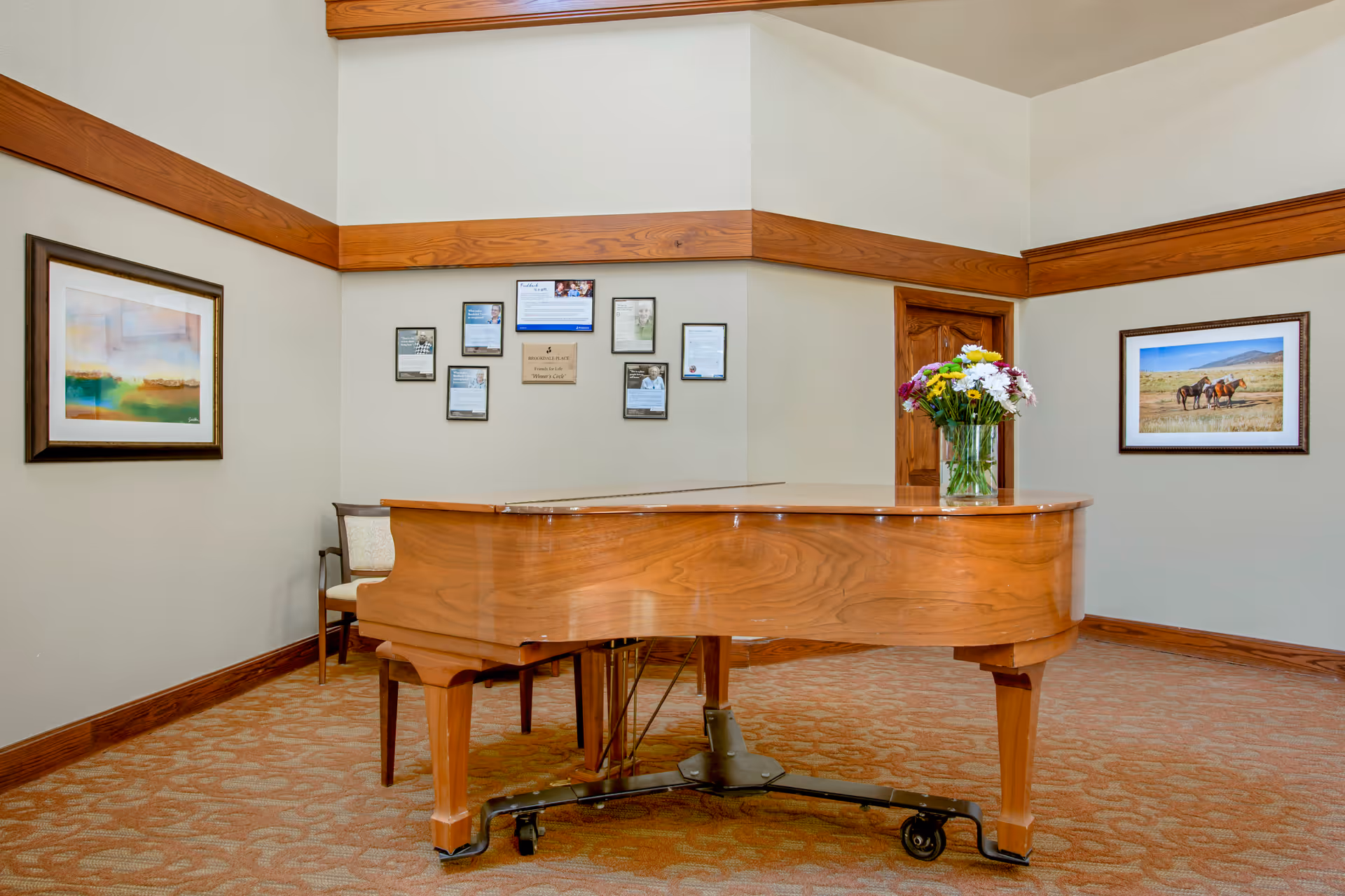 A wooden grand piano on wheels is placed in a carpeted room with beige walls and wooden trim. A vase with colorful flowers sits on top of the piano. The walls are decorated with framed pictures and certificates. There is a wooden door and a chair partially visible behind the piano.