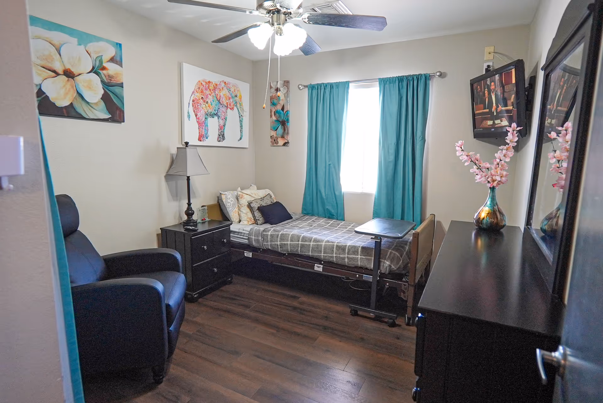 A small bedroom with a single bed covered with a gray checkered blanket and multiple pillows. Next to the bed is a black nightstand with a lamp and a remote control. A black recliner chair is positioned near the entrance. The room has wooden flooring, teal curtains covering a window, and three colorful wall art pieces including a floral painting and a multicolored elephant. A flat-screen TV is mounted on the wall opposite the bed, above a black dresser with a vase of pink flowers. A ceiling fan with lights is on the ceiling.