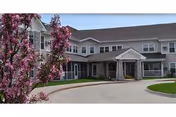 Front entrance of a two-story senior living building with a circular driveway and a flowering tree in the foreground.