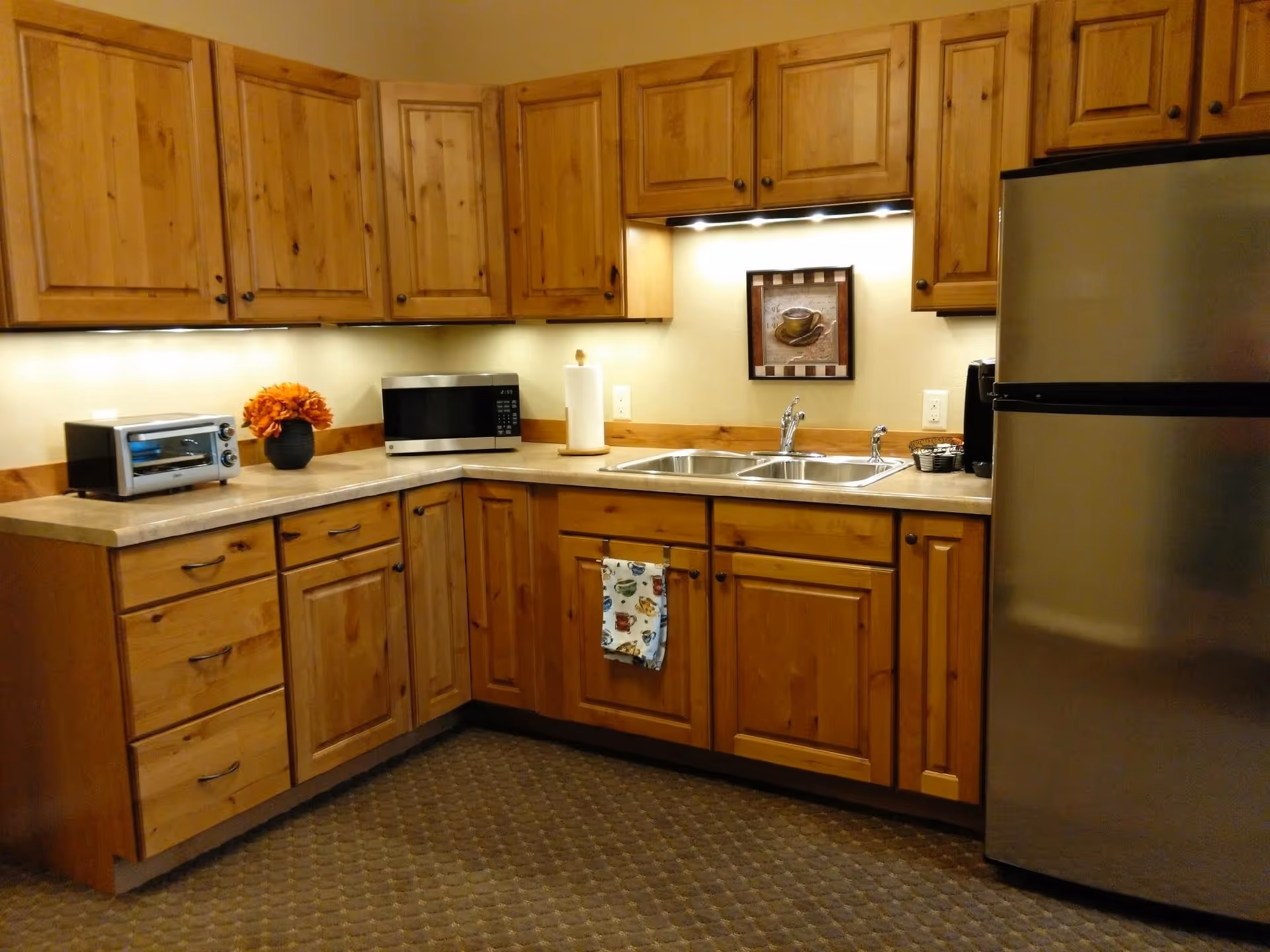 A kitchen with wooden cabinets, a stainless steel refrigerator, a double sink, a microwave, a toaster oven, a paper towel holder, a coffee maker, and a small vase with orange flowers on the countertop. There is a framed picture of a coffee cup on the wall above the sink.