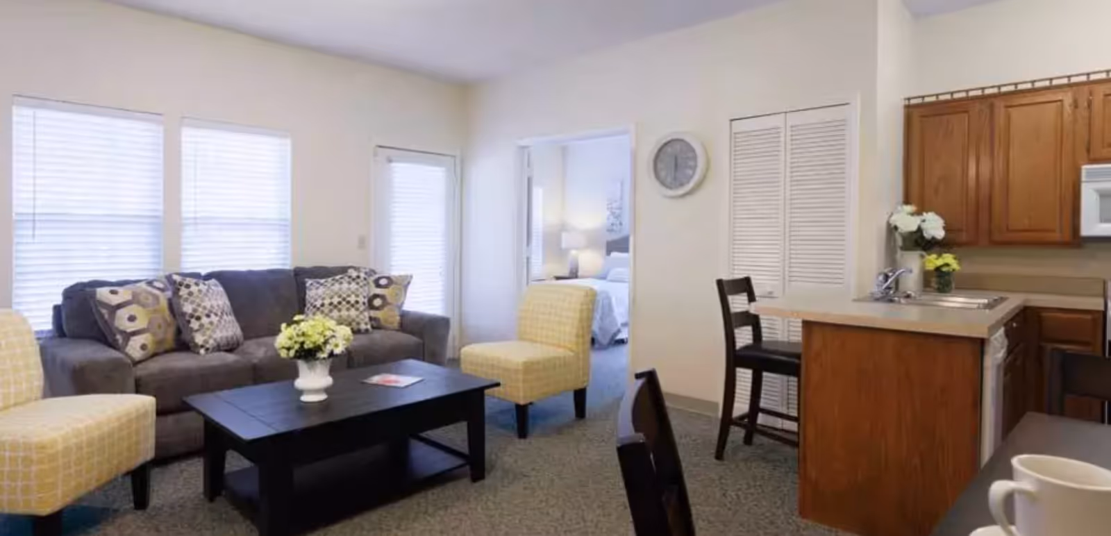 Bright living room with a sofa, patterned armchairs, a coffee table, and a kitchenette with bar seating, with a bedroom visible through a doorway.