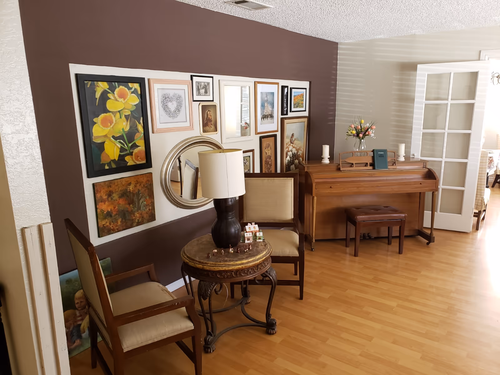 A cozy living area with two chairs and a small round table with a lamp in front of a gallery wall, and an upright piano to the right.